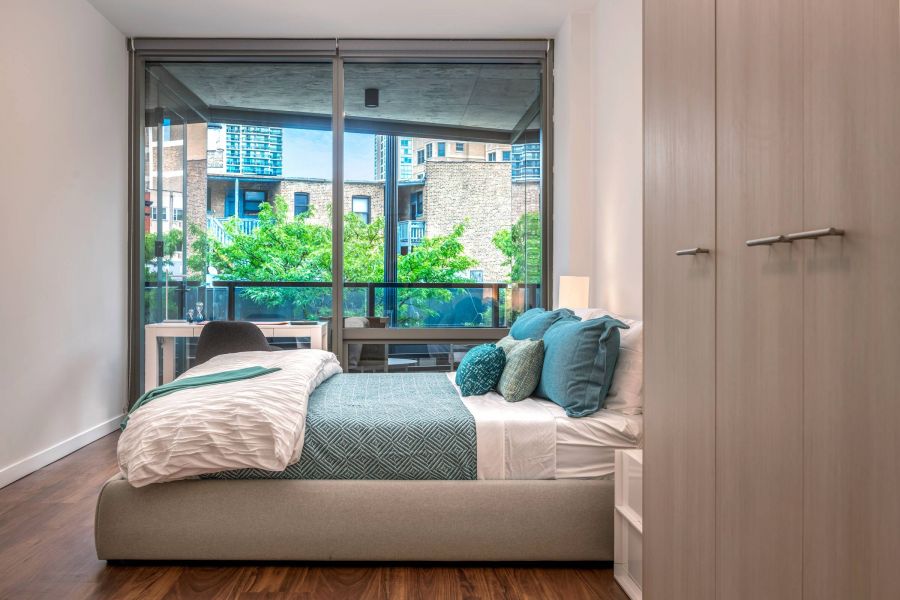 Optima Lakeview® Apartments in Chicago, IL Modern bedroom with a large window, city view, teal bedding, and wooden floors.