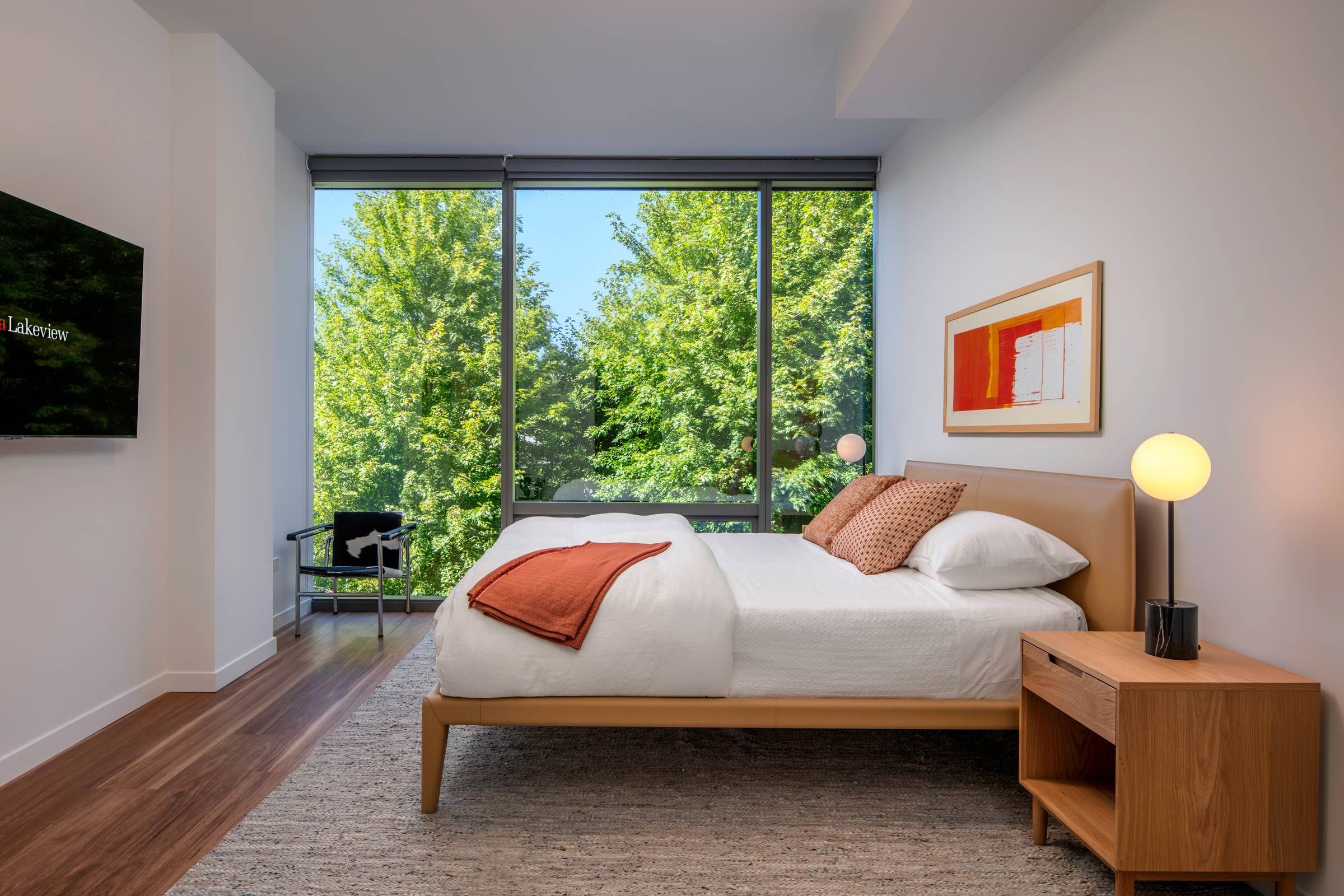 Optima Lakeview® Apartments in Chicago, IL Modern bedroom with large windows, a wooden bed, orange accents, and a view of green trees outside.