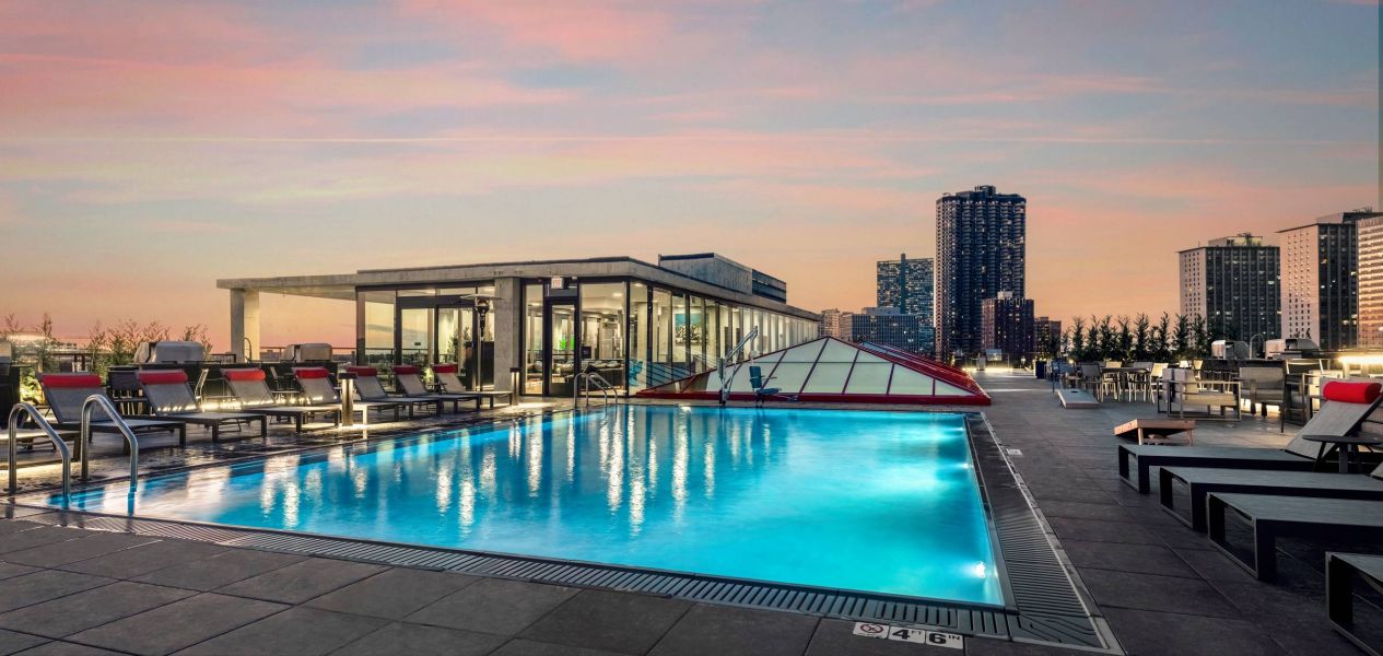 Optima Lakeview® Apartments in Chicago, IL Rooftop pool at sunset with lounge chairs, city skyline, and modern glass building in the background.