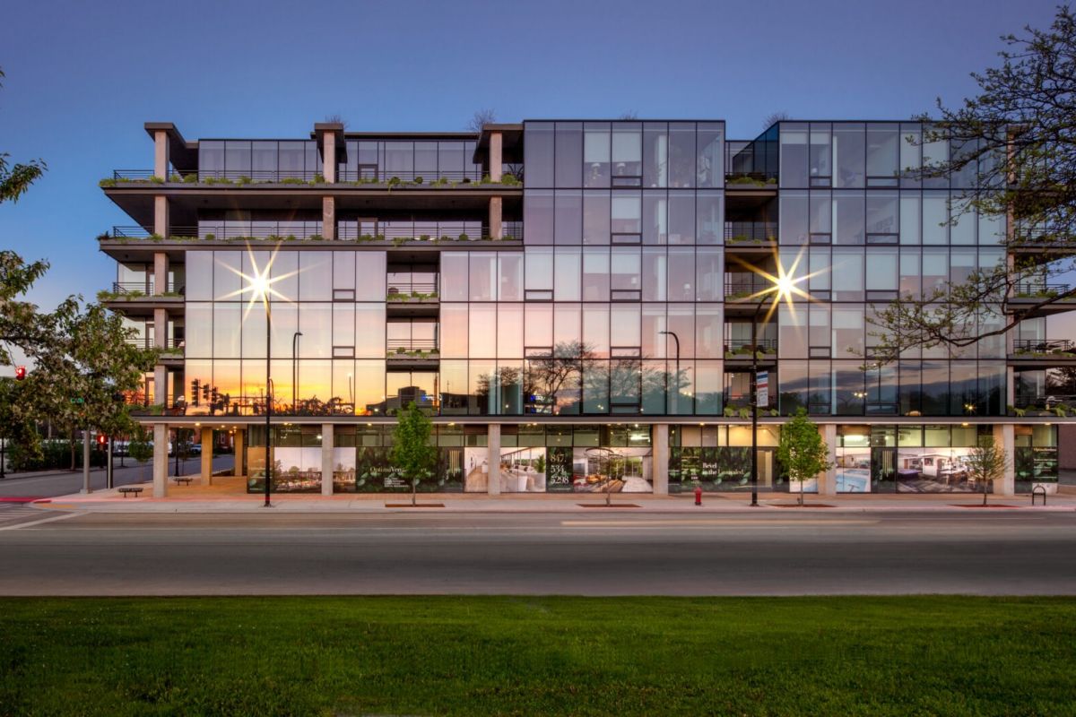 Optima Lakeview® Apartments in Chicago, IL Modern glass apartment building reflecting sunset, showcasing stunning architecture and abundant natural light, with trees and street in front.