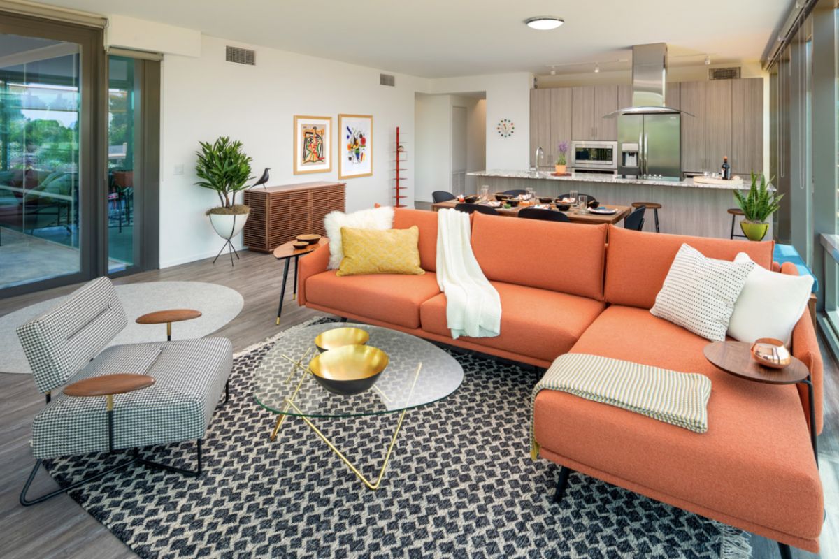 Optima Lakeview® Apartments in Chicago, IL Modern living room with orange sectional sofa, patterned rug, and a glass coffee table creates a vibrant mood, with open kitchen in the space.