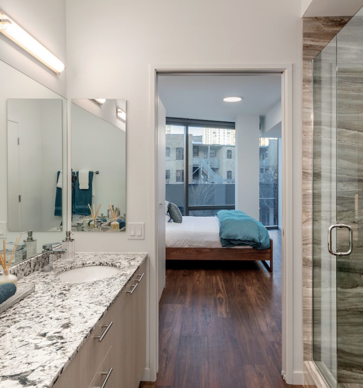 Optima Lakeview® Apartments in Chicago, IL Modern bathroom decor with double sinks and a glass shower, leading to a bedroom with large windows and wood flooring.