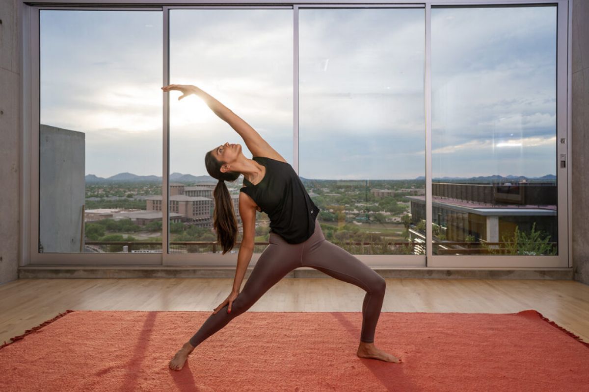 Optima Lakeview® Apartments in Chicago, IL Woman practicing yoga and mindfulness in a modern room with large windows, cityscape and mountains visible at sunset.