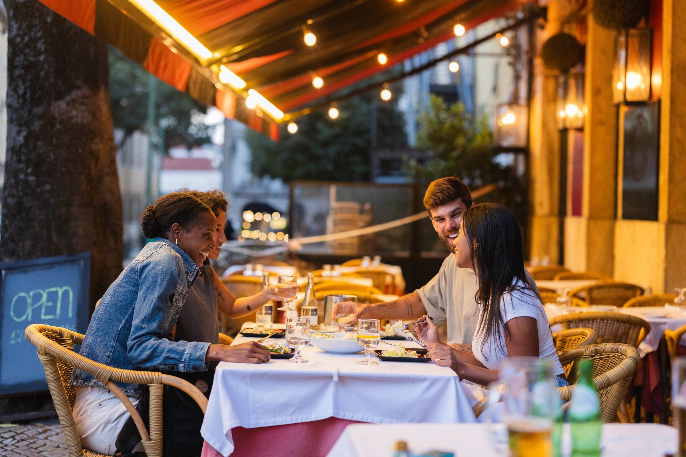Optima Lakeview® Apartments in Chicago, IL Four friends laugh and dine together at an outdoor restaurant in the evening under string lights.