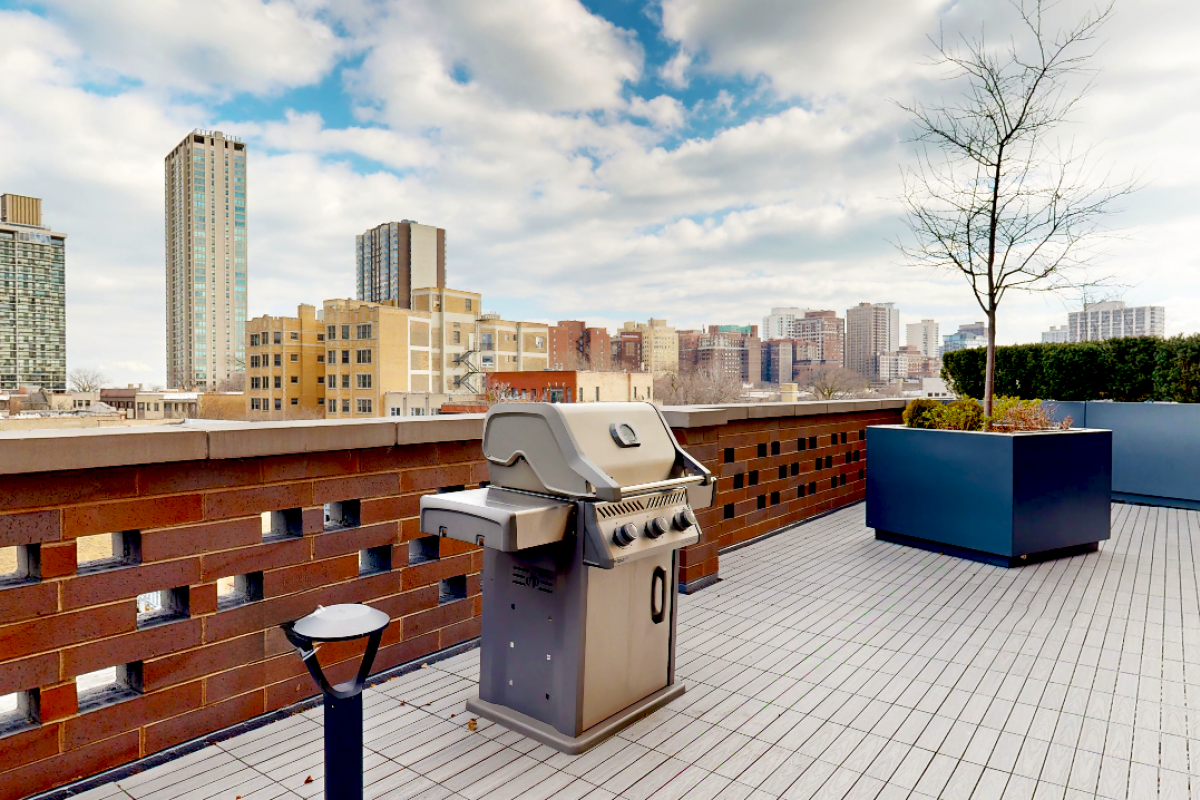 Optima Lakeview® Apartments in Chicago, IL Rooftop patio at Optima with a grill, potted plants, and city buildings—a perfect outdoor space under a partly cloudy sky.