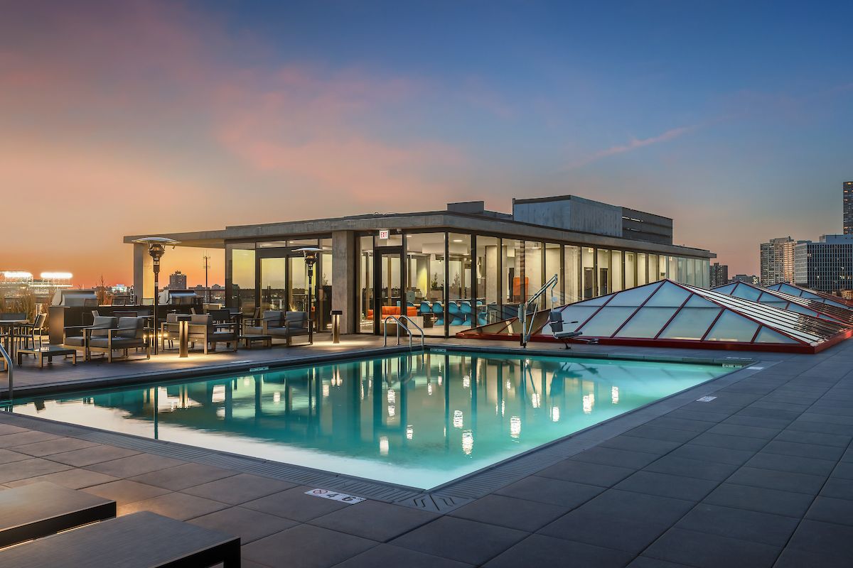 Optima Lakeview® Apartments in Chicago, IL Rooftop pool and lounge area with city skyline views at sunset, alfresco cooking spaces, and modern glass building by Optima Communities.