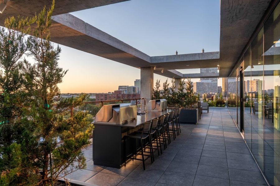 Optima Lakeview® Apartments in Chicago, IL Modern rooftop patio with grills, bar seating, greenery, and city skyline views at sunset.