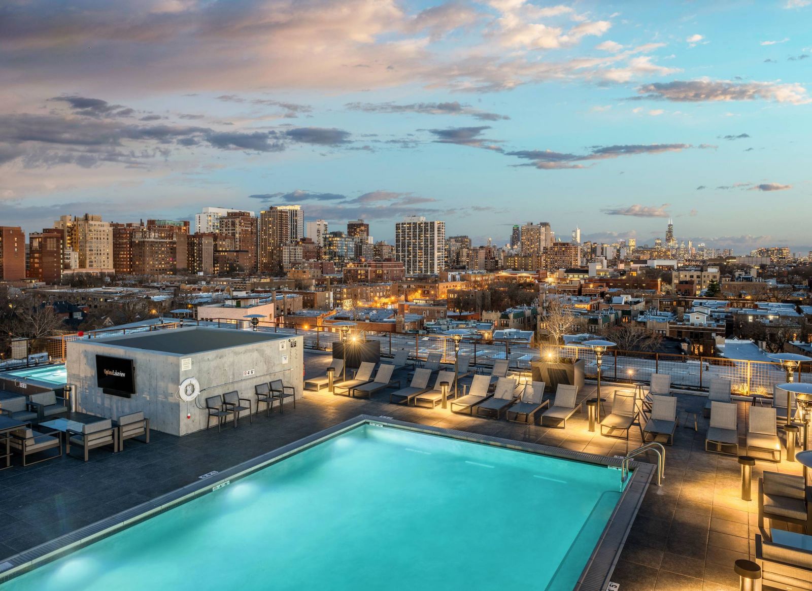 Optima Lakeview® Apartments in Chicago, IL Rooftop pool with lounge chairs overlooking a city skyline at sunset, under a partly cloudy sky.