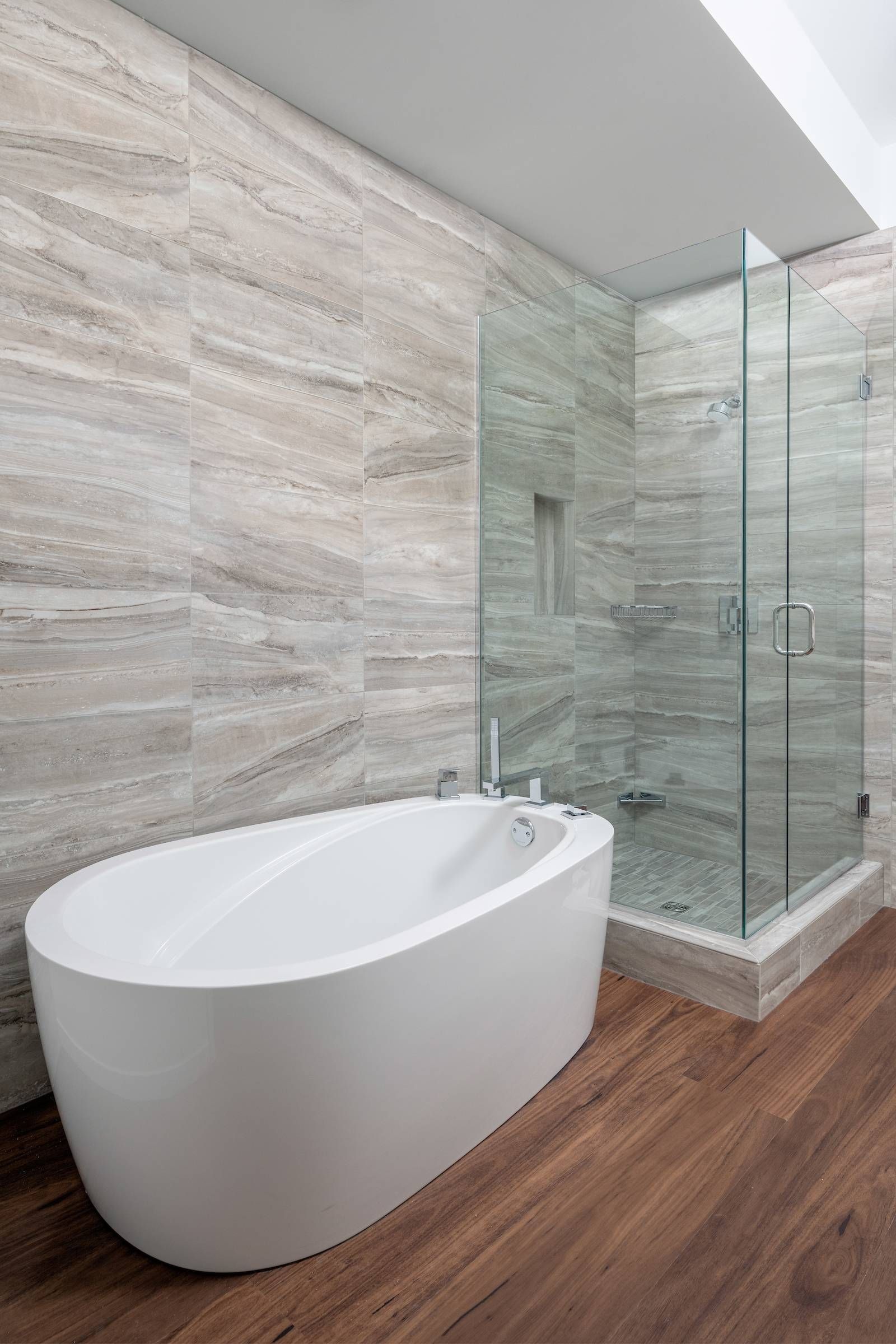 Optima Lakeview® Apartments in Chicago, IL Modern bathroom with a white oval bathtub, glass shower, wood floor, and beige marble walls.
