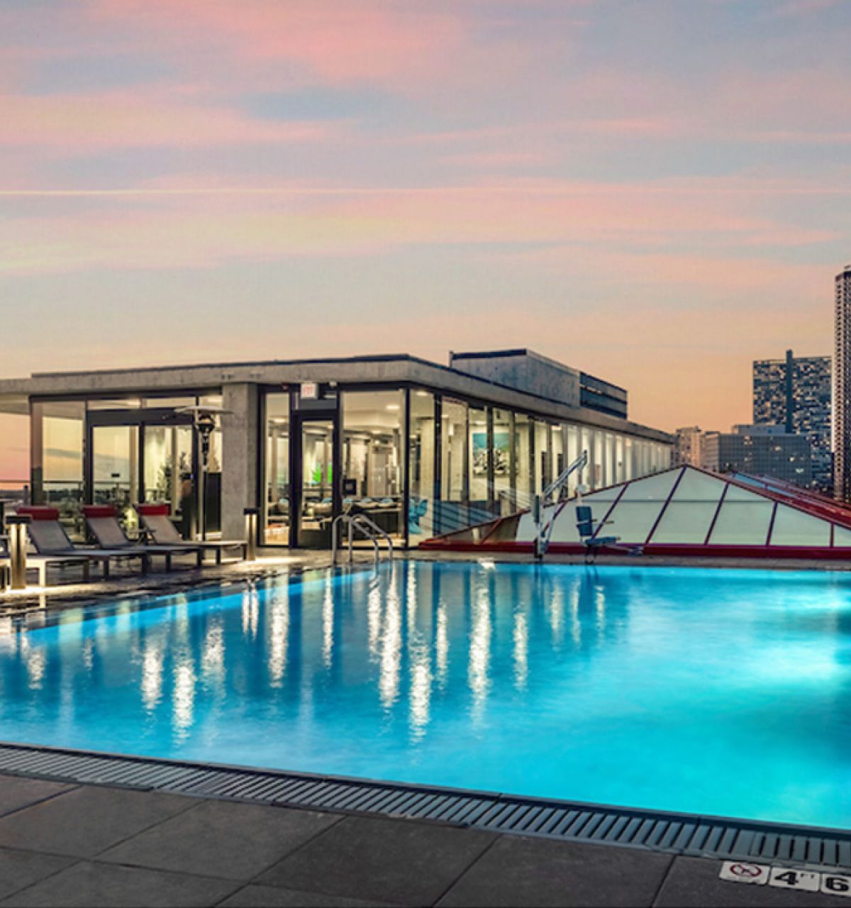 Optima Lakeview® Apartments in Chicago, IL Rooftop pool at sunset at Optima Lakeview, with lounge chairs, city skyline, glass building, and tall buildings in the background.