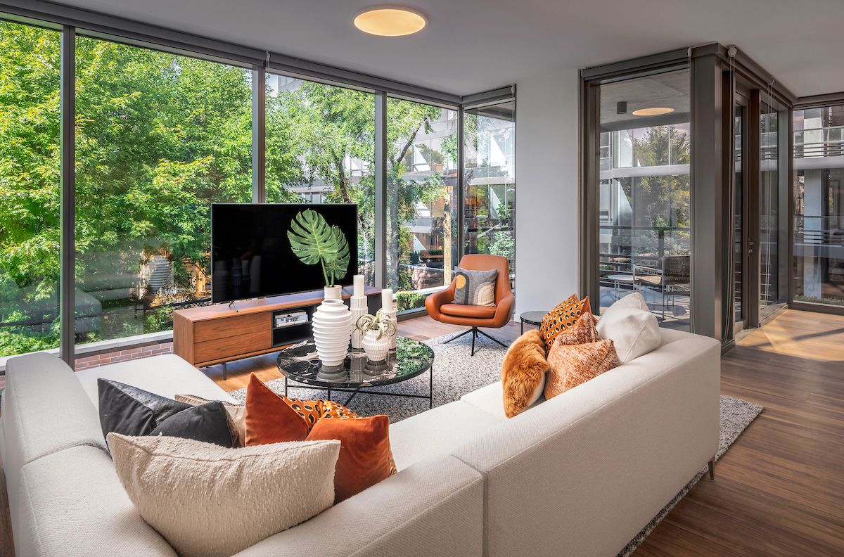 Optima Lakeview® Apartments in Chicago, IL Modern living room with large windows, neutral sofa, orange chair, and TV, overlooking green trees outside.