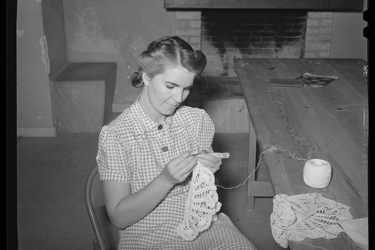 Optima Lakeview® Apartments in Chicago, IL Woman in gingham dress sits crocheting lace at a wooden table near a fireplace at Firefly Fiber Arts Studio.