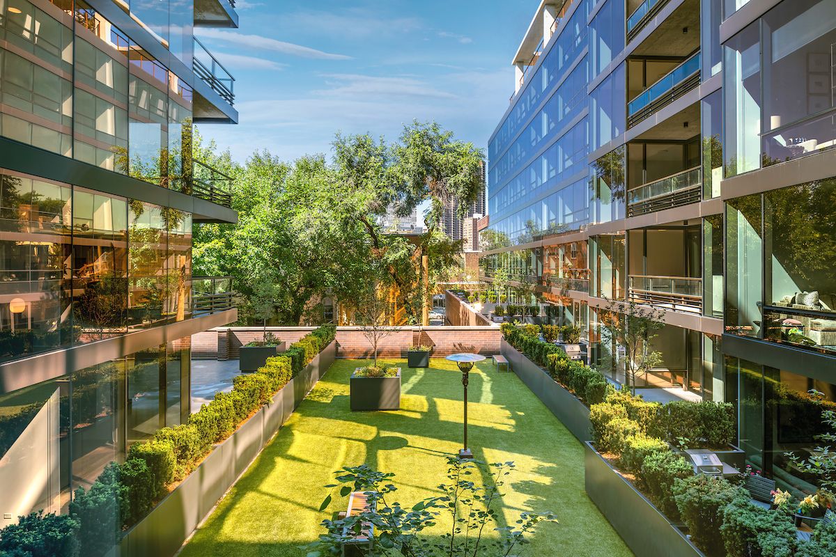 Optima Lakeview® Apartments in Chicago, IL Modern glass buildings with balconies at Optima Lakeview overlook a pet-friendly green courtyard garden with trees and shrubs on a sunny day.