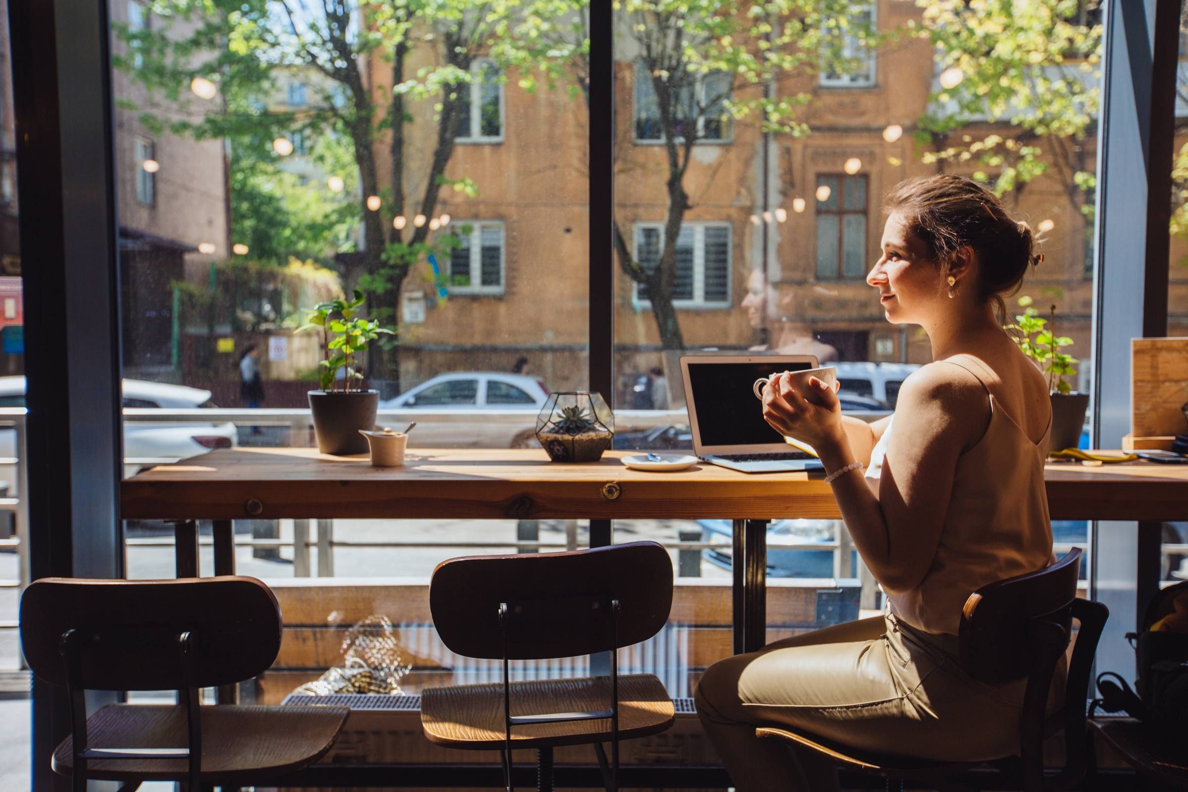 Optima Lakeview® Apartments in Chicago, IL Woman sitting at a cafe window with a coffee cup, laptop, and sunlight streaming through large windows.