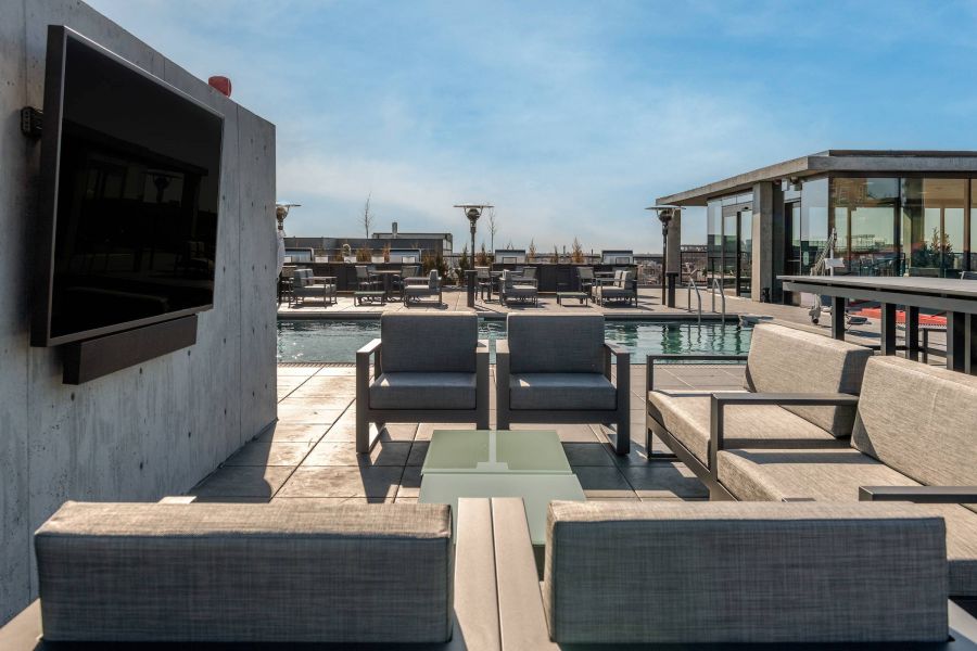 Optima Lakeview® Apartments in Chicago, IL Modern rooftop lounge with gray seating, a TV, and a pool under a clear blue sky.