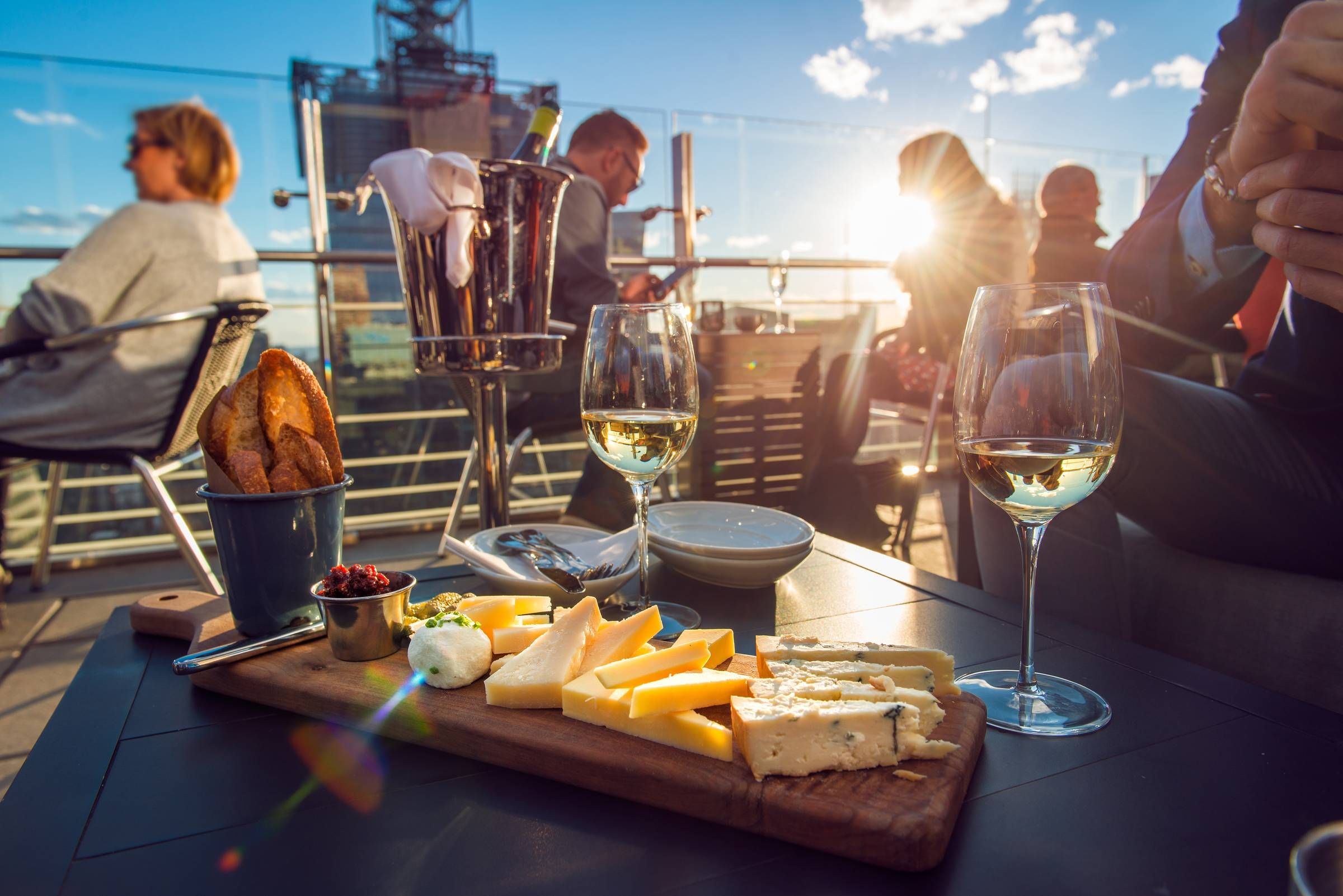 Optima Lakeview® Apartments in Chicago, IL Cheese platter, bread, and wine on a rooftop patio at sunset with people seated in the background.