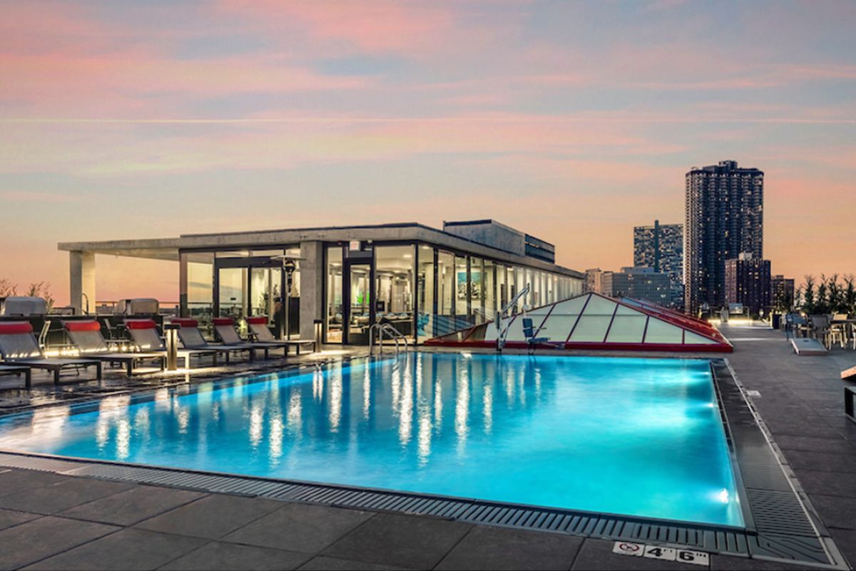 Optima Lakeview® Apartments in Chicago, IL Rooftop pool at sunset at Optima Lakeview, with lounge chairs, city skyline, glass building, and tall buildings in the background.