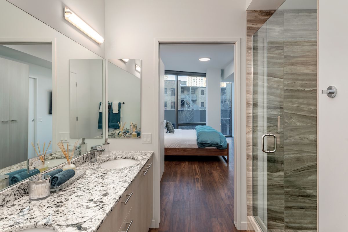 Optima Lakeview® Apartments in Chicago, IL Modern bathroom decor with double sinks and a glass shower, leading to a bedroom with large windows and wood flooring.