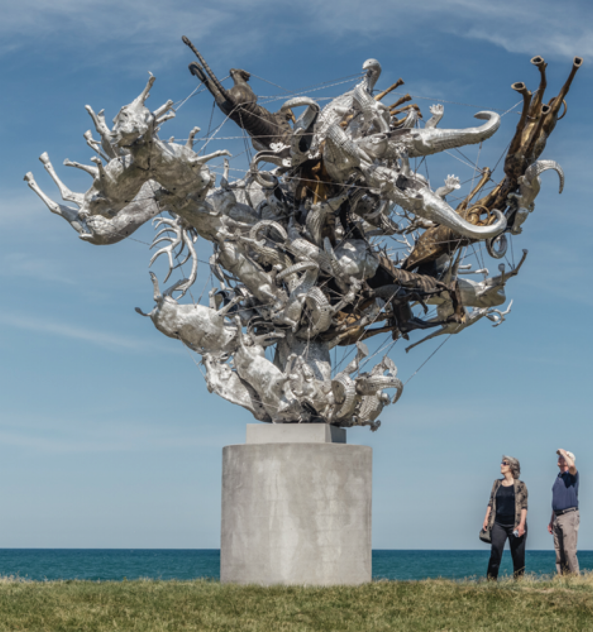 Optima Lakeview® Apartments in Chicago, IL Two people stand near Agrifolia Majoris, a large abstract metallic sculpture, part of Chicago Public Art, by the ocean on a grassy area.