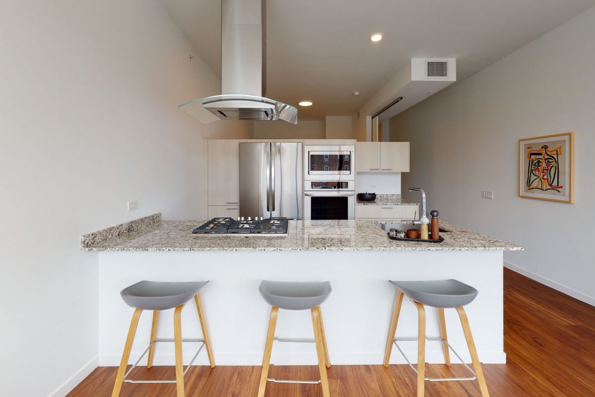 Optima Lakeview® Apartments in Chicago, IL Modern kitchen in Optima Lakeview’s 1B-01 residences features a granite island, three gray stools, and stainless steel appliances.