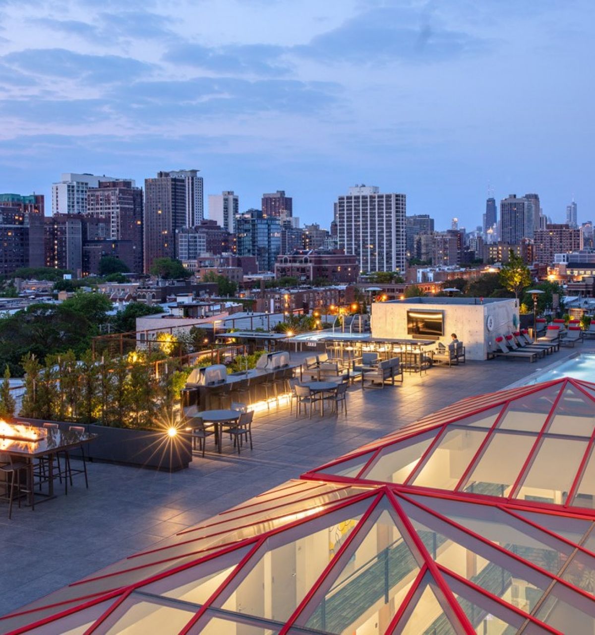 Optima Lakeview® Apartments in Chicago, IL Rooftop lounge at Optima Lakeview with fire pits, tables, and a pool overlooking the city skyline at dusk.