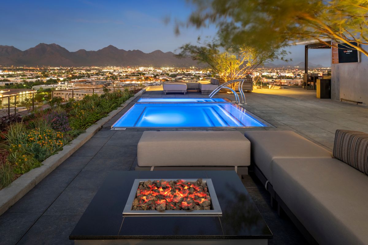 Optima Lakeview® Apartments in Chicago, IL A modern rooftop pool and fire pit lounge offer good neighbor vibes, overlooking a cityscape with mountains at sunset.