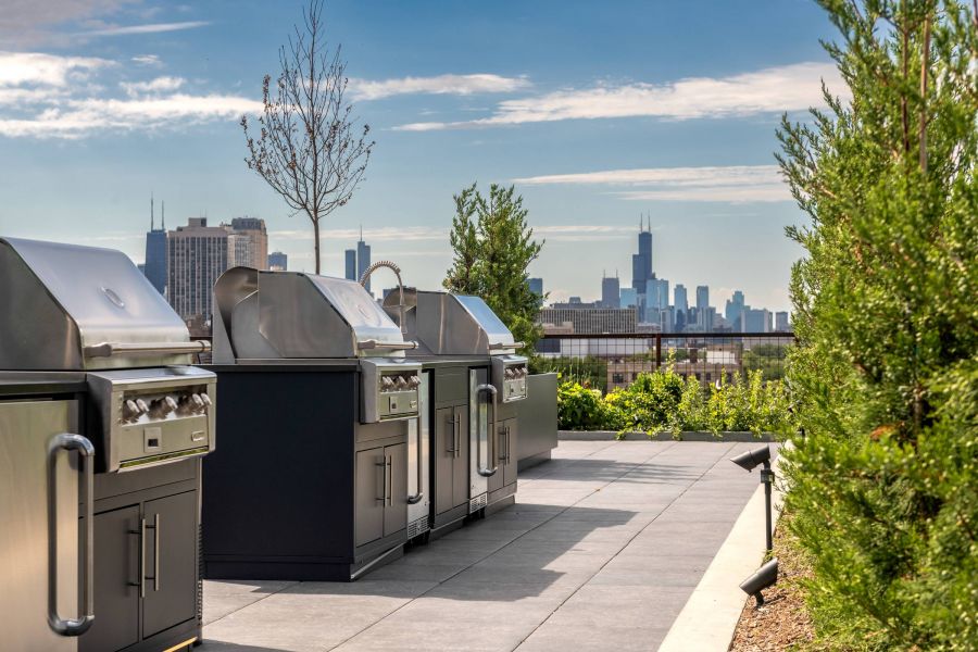 Optima Lakeview® Apartments in Chicago, IL Three stainless steel grills on a rooftop patio with a city skyline in the background under a blue sky.