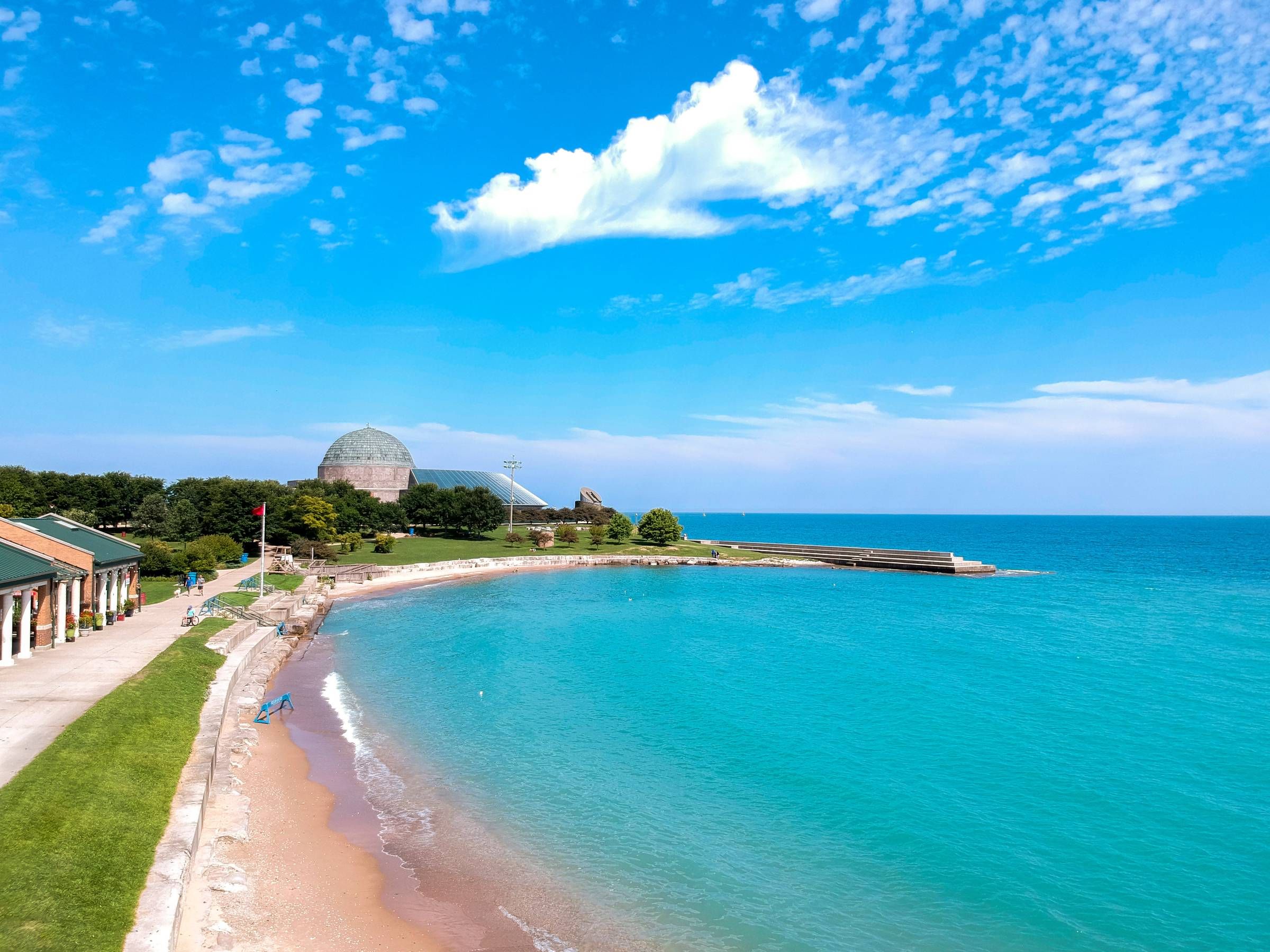 Optima Lakeview® Apartments in Chicago, IL A bright blue lake curves around a sandy beach with a domed building and green trees in the background.