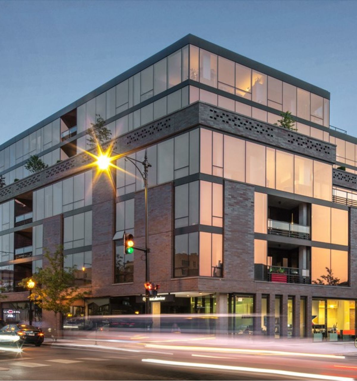 Optima Lakeview® Apartments in Chicago, IL Modern glass and brick apartment building on a busy city street at sunset with light trails from cars.