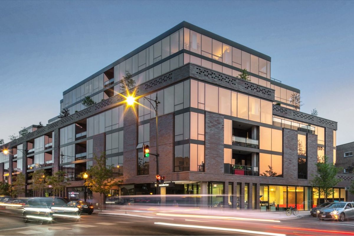 Optima Lakeview® Apartments in Chicago, IL Modern glass and brick apartment building on a busy city street at dusk, with blurred car lights passing by.