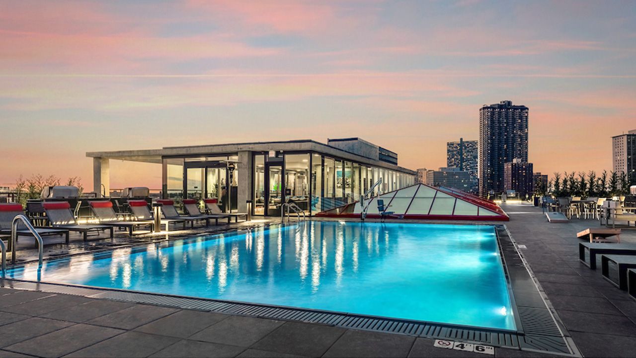 Optima Lakeview® Apartments in Chicago, IL Rooftop pool at sunset at Optima Lakeview, with lounge chairs, city skyline, glass building, and tall buildings in the background.