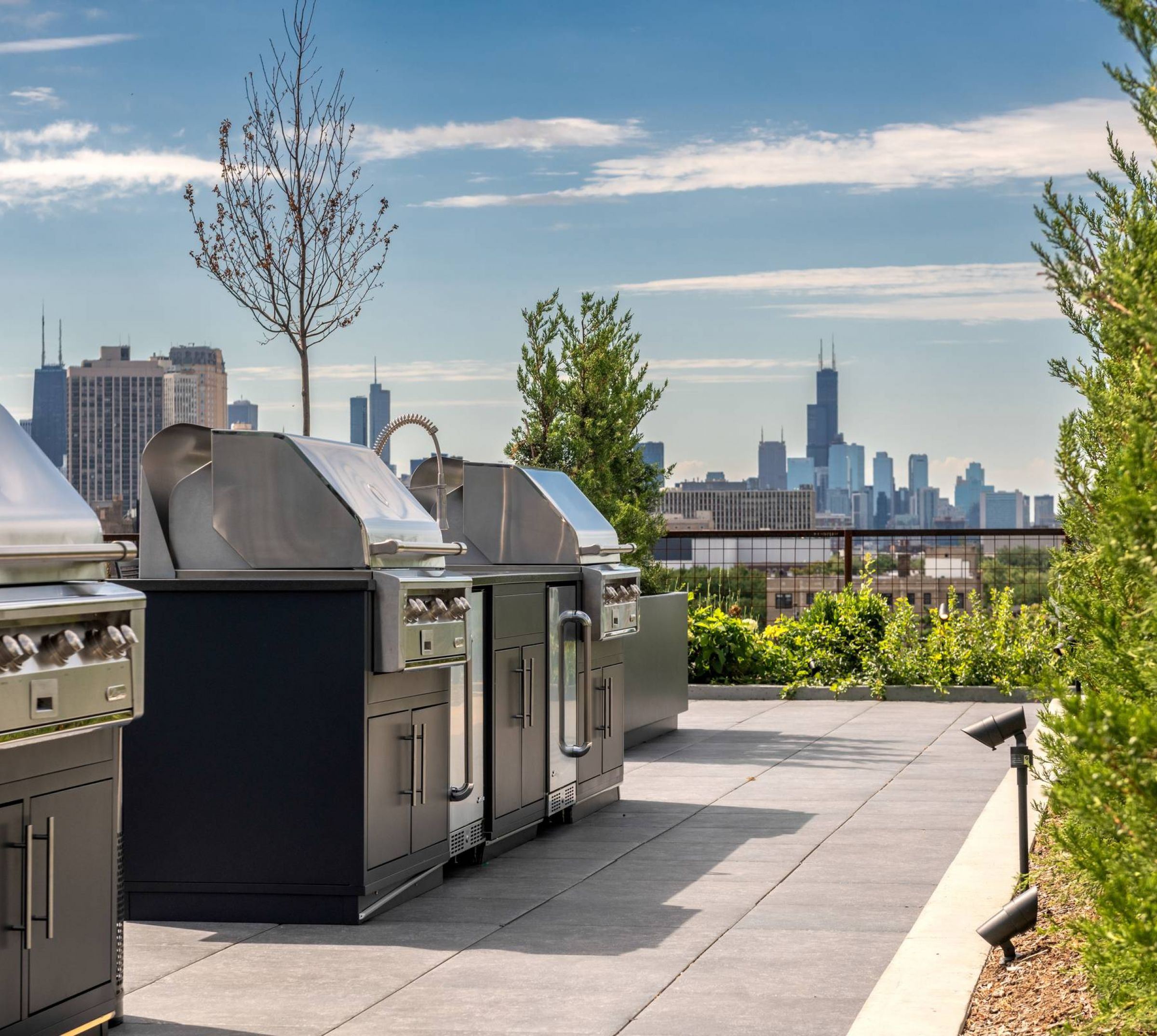 Optima Lakeview® Apartments in Chicago, IL Three stainless steel grills on a rooftop patio with a city skyline in the background under a blue sky.