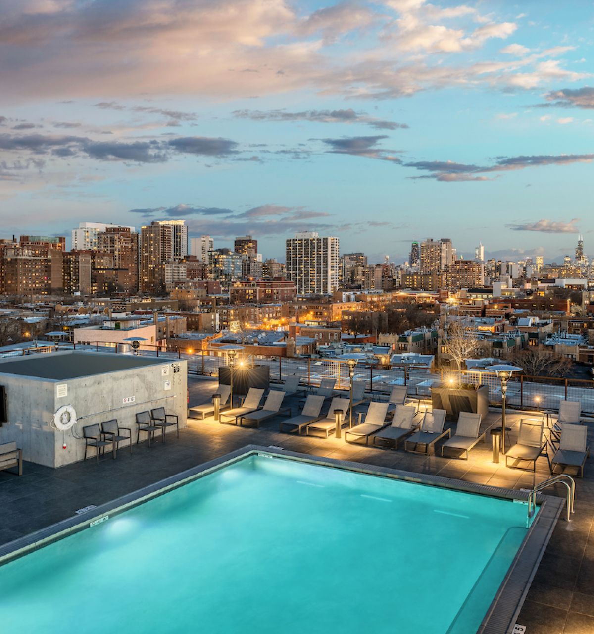 Optima Lakeview® Apartments in Chicago, IL Rooftop pool with lounge chairs overlooking a city skyline at sunset, under a partly cloudy sky.