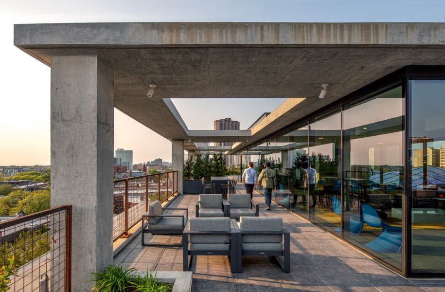 Optima Lakeview® Apartments in Chicago, IL Modern rooftop terrace with seating, greenery, and people walking, overlooking a city skyline at sunset.