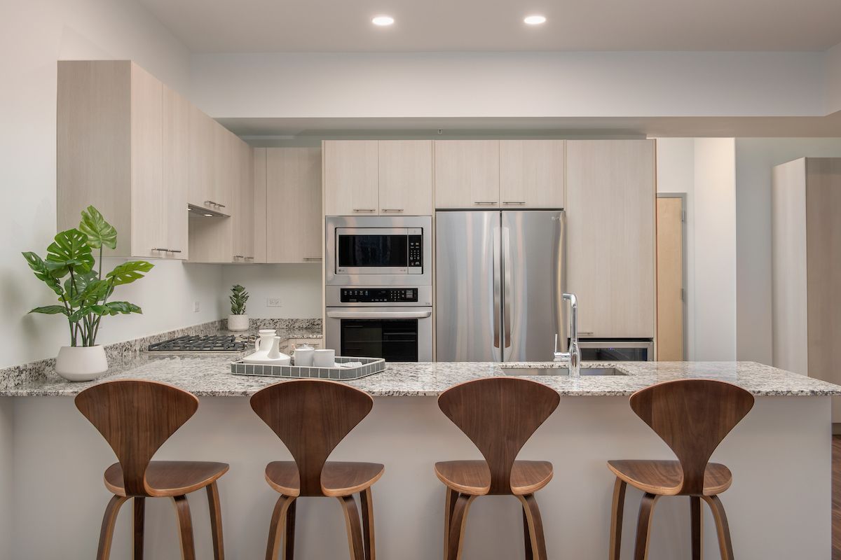 Optima Lakeview® Apartments in Chicago, IL Modern kitchen design with light wood cabinets, stainless steel appliances, and four wooden barstools at an Optima granite island.