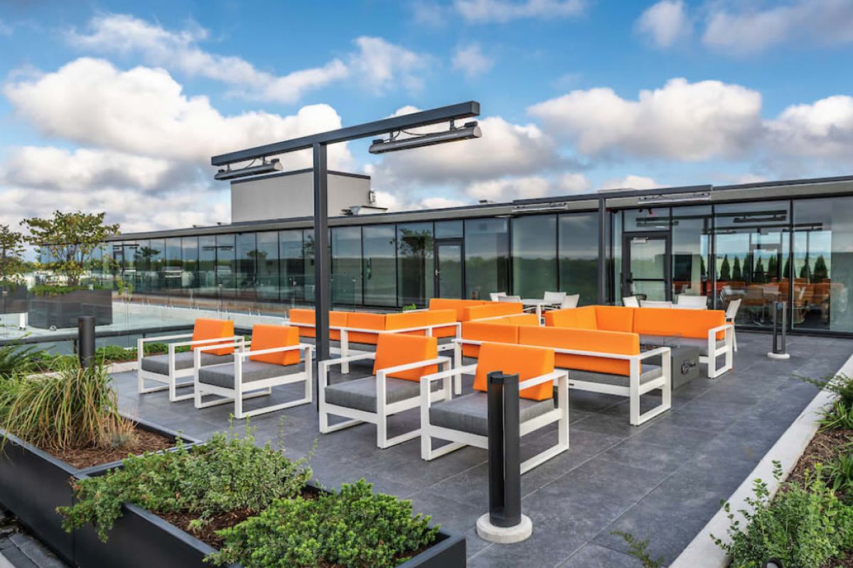 Optima Lakeview® Apartments in Chicago, IL Modern rooftop patio with orange seating, greenery, and glass-walled building showcases the benefits of green roofs under a partly cloudy sky.