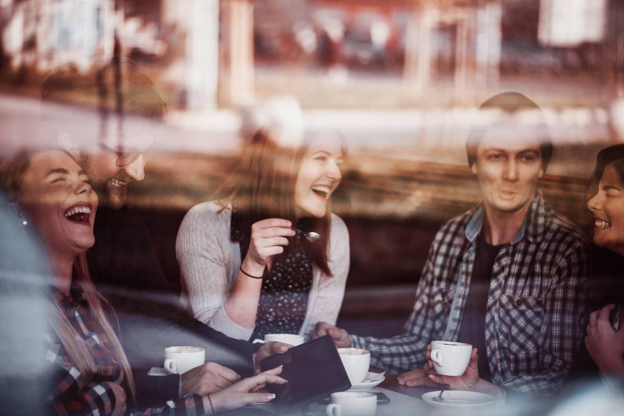 Optima Lakeview® Apartments in Chicago, IL Five friends sit at a café table, laughing and drinking coffee, seen through a window.