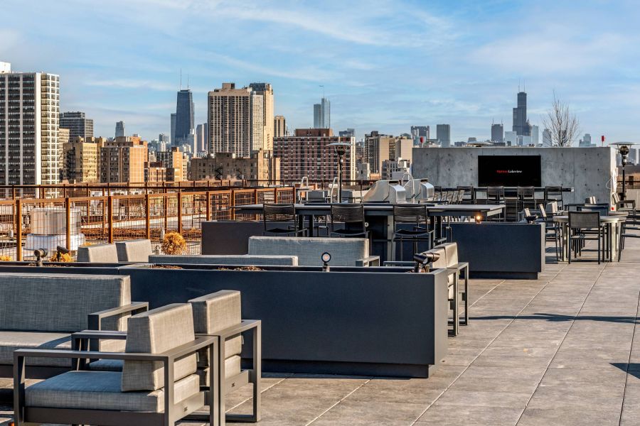 Optima Lakeview® Apartments in Chicago, IL Modern rooftop lounge with gray seating, city skyline, and tall buildings in the background under a blue sky.