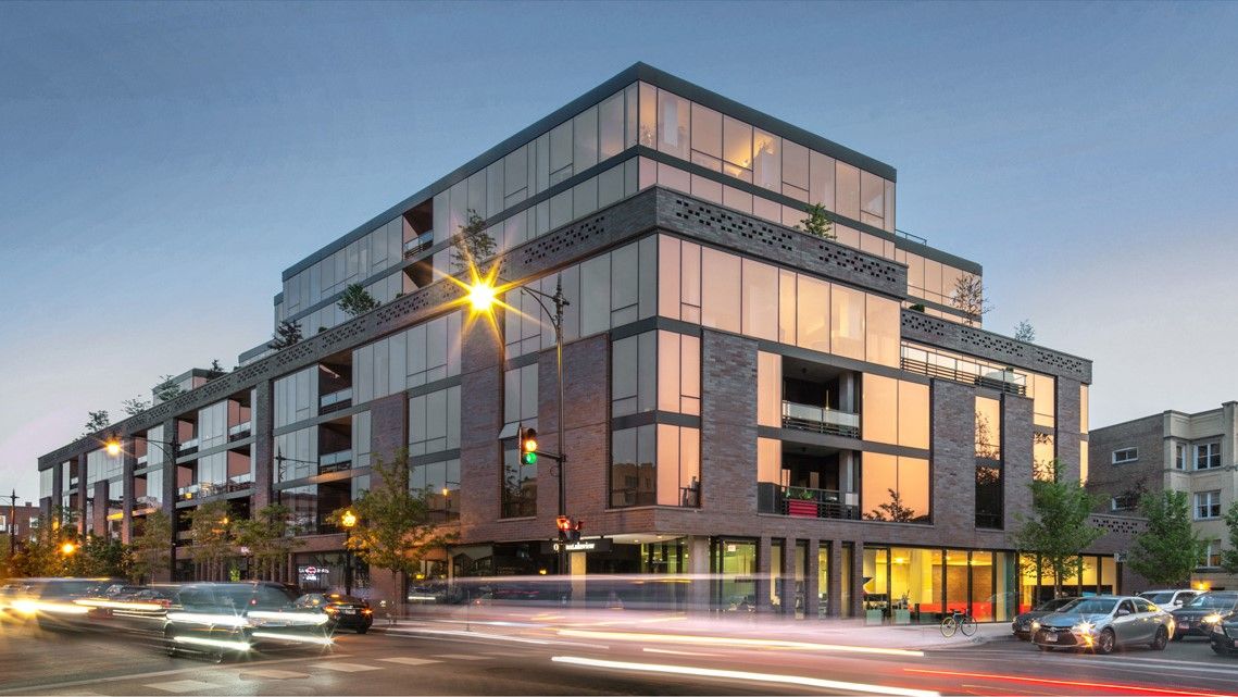 Optima Lakeview® Apartments in Chicago, IL A multi-story Optima Lakeview building with a brick and glass facade at a street corner during twilight, featuring blurred car light trails.