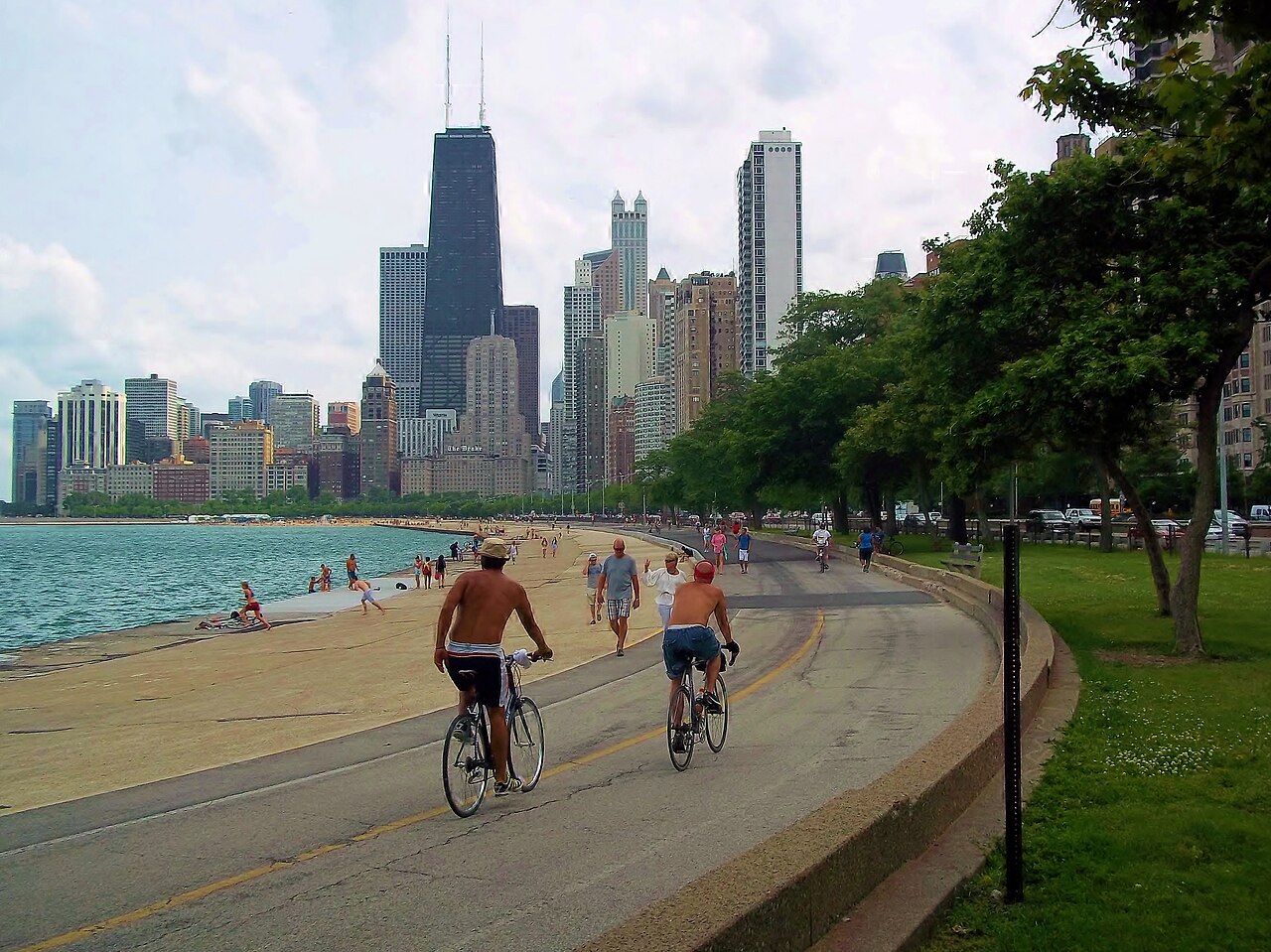 Optima Lakeview® Apartments in Chicago, IL People biking and walking along a lakeside path in the Chicago Lakeview neighborhood, with the skyline and lakeside cafés in the background.