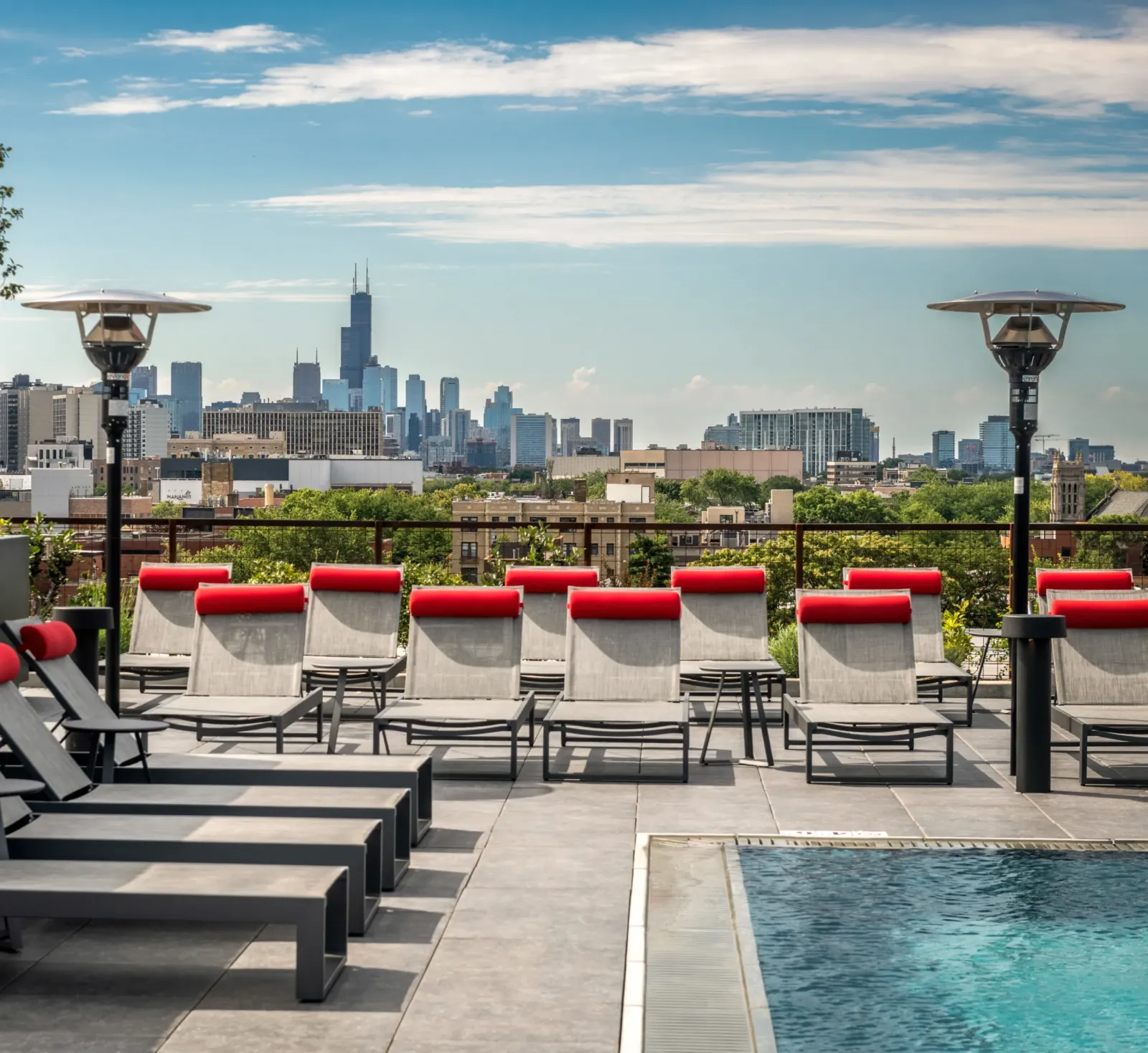Optima Lakeview® Apartments in Chicago, IL Rooftop pool with lounge chairs and heaters, city skyline and skyscrapers in the background under a blue sky.