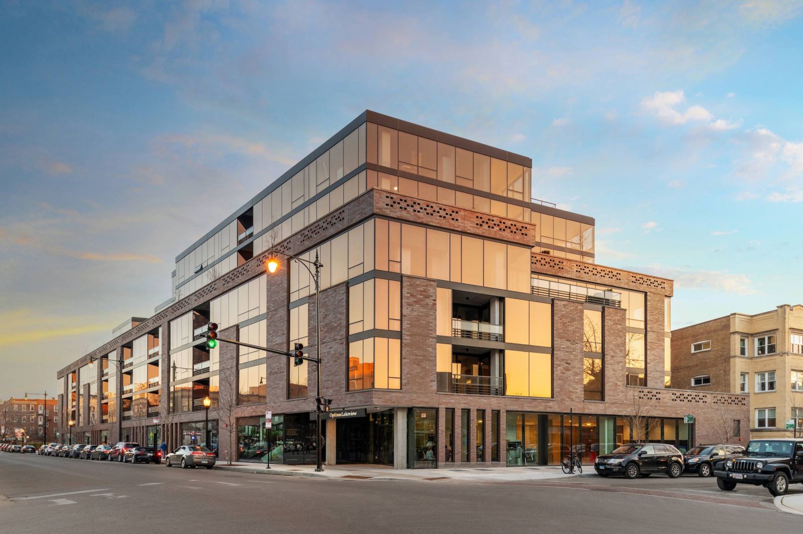 Optima Lakeview® Apartments in Chicago, IL Modern multi-story building with large glass windows on a city street at sunset, reflecting the sky.