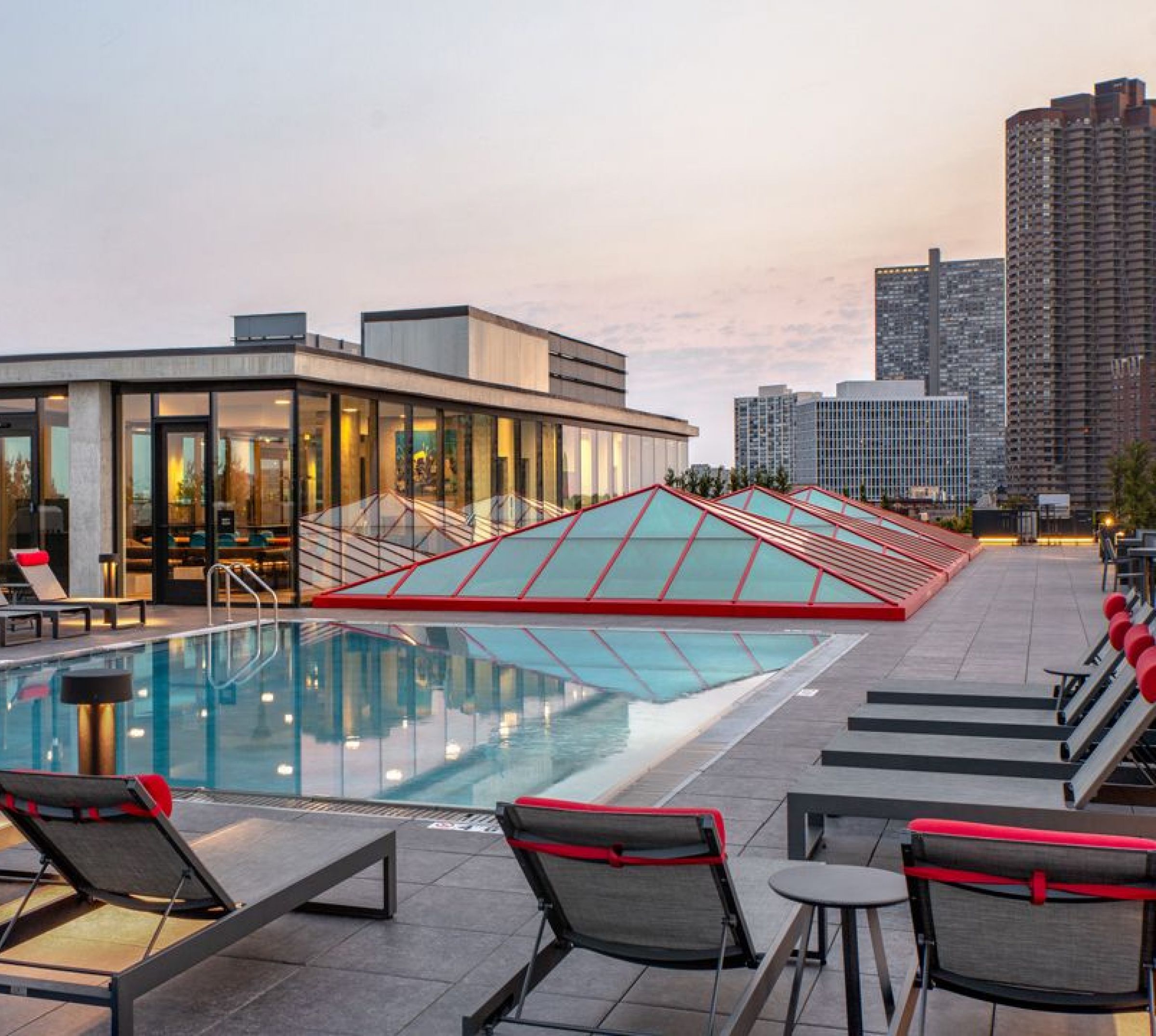 Optima Lakeview® Apartments in Chicago, IL Rooftop pool with red-accented lounge chairs, modern building, and city skyline in the background at sunset.
