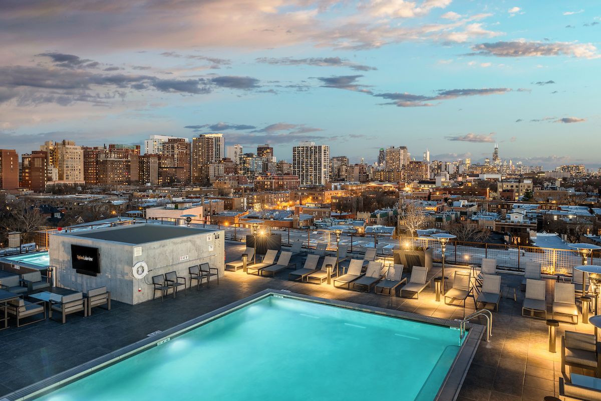 Optima Lakeview® Apartments in Chicago, IL Rooftop pool with lounge chairs overlooking a city skyline at sunset, under a partly cloudy sky.