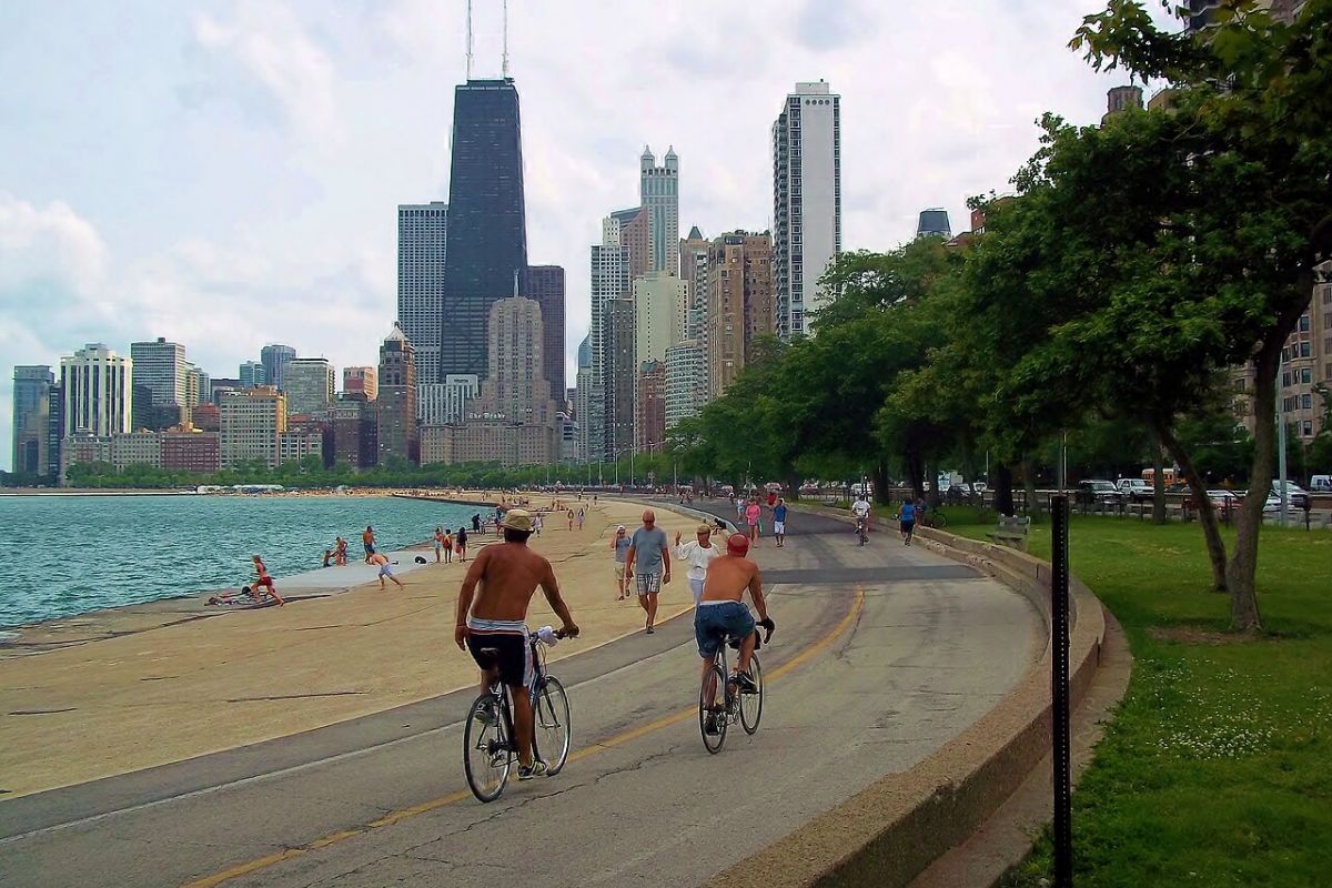 Optima Lakeview® Apartments in Chicago, IL People biking and walking along a lakeside path in the Chicago Lakeview neighborhood, with the skyline and lakeside cafés in the background.