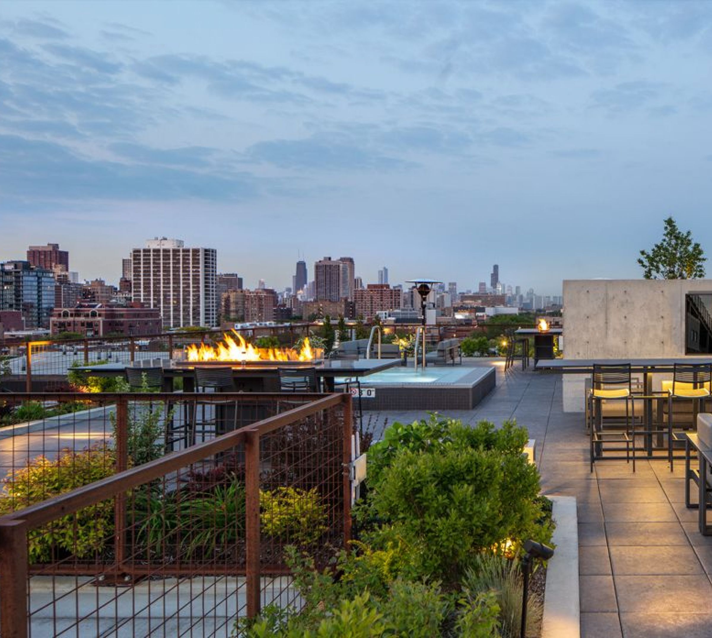 Optima Lakeview® Apartments in Chicago, IL Rooftop terrace with fire pit, seating, greenery, and city skyline view at dusk.
