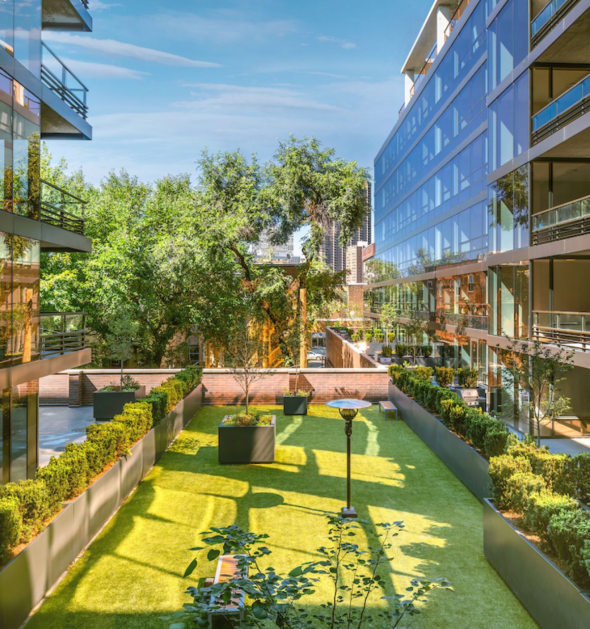 Optima Lakeview® Apartments in Chicago, IL Modern glass buildings with balconies at Optima Lakeview overlook a pet-friendly green courtyard garden with trees and shrubs on a sunny day.