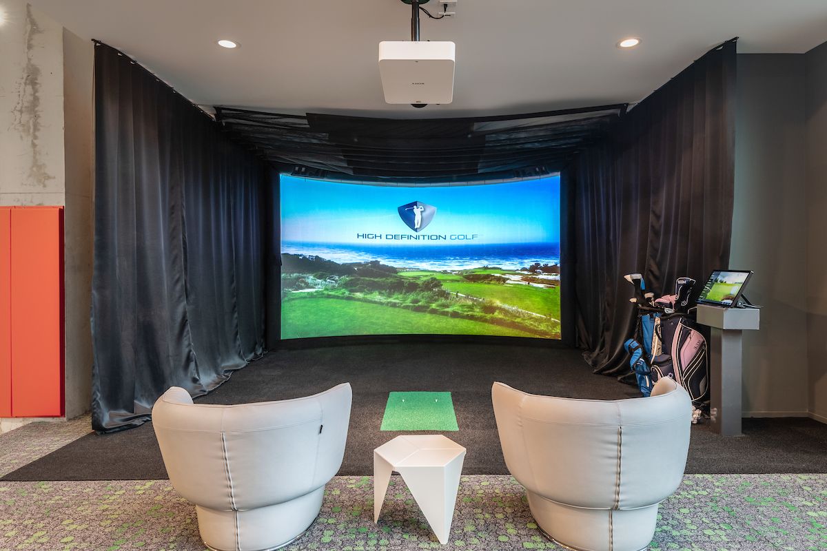 Optima Lakeview® Apartments in Chicago, IL Two gray chairs face a golf simulator screen, perfect for indoor golf, with clubs on the right and a small white table in between.