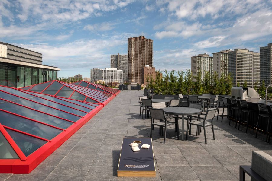 Optima Lakeview® Apartments in Chicago, IL Rooftop patio with tables, chairs, greenery, city skyline views, and a cornhole game under a partly cloudy sky.