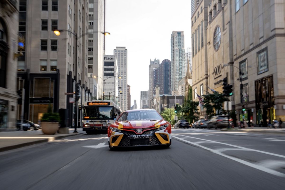 Optima Lakeview® Apartments in Chicago, IL A NASCAR race car speeds down a Chicago street, with tall buildings and a city bus in the background.